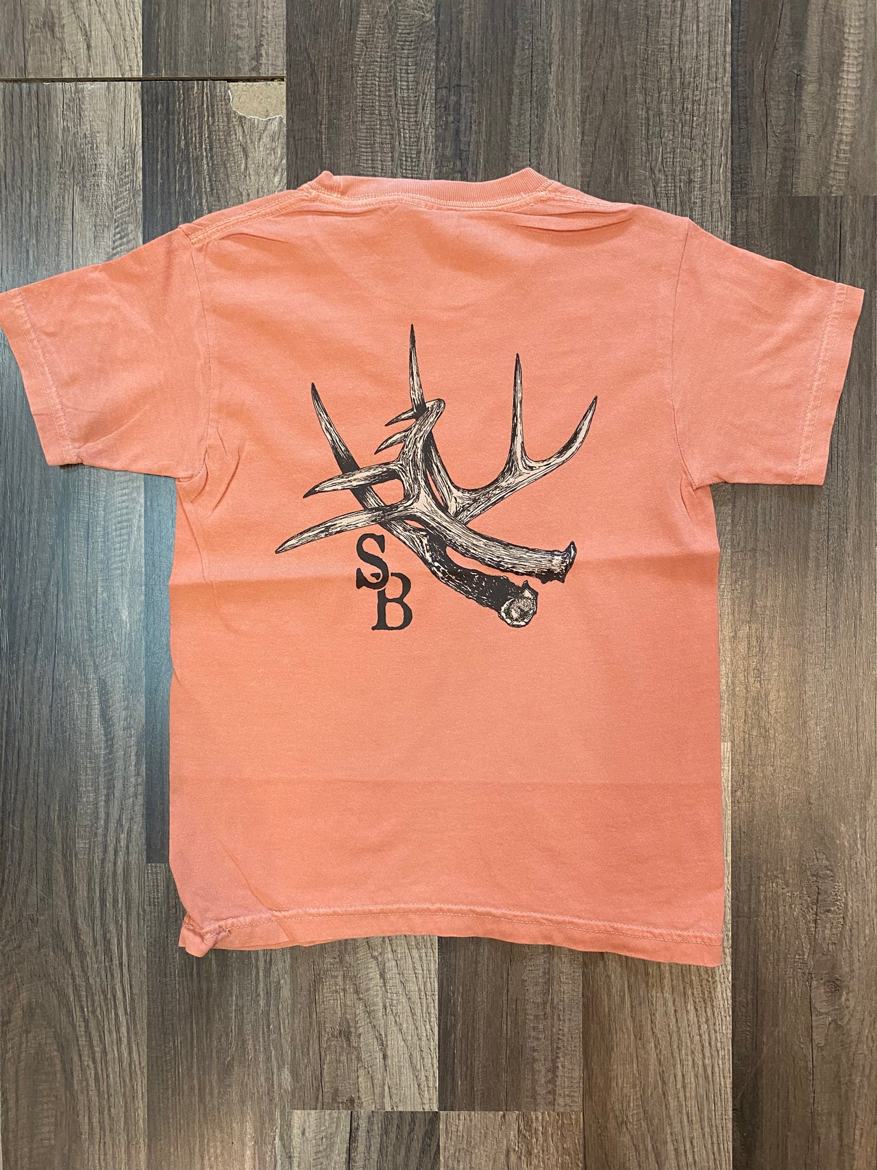 "Image of a Youth Speckle Bellies Terracotta Color Antler Short Sleeve Tee in Gildan Natural, featuring a soft washed garment dyed fabric with twill-taped neck and shoulders. The tee has double-needle stitching on the collar, armhole, sleeves, and bottom hem. Pigment shades naturally vary, creating a unique and textured appearance."