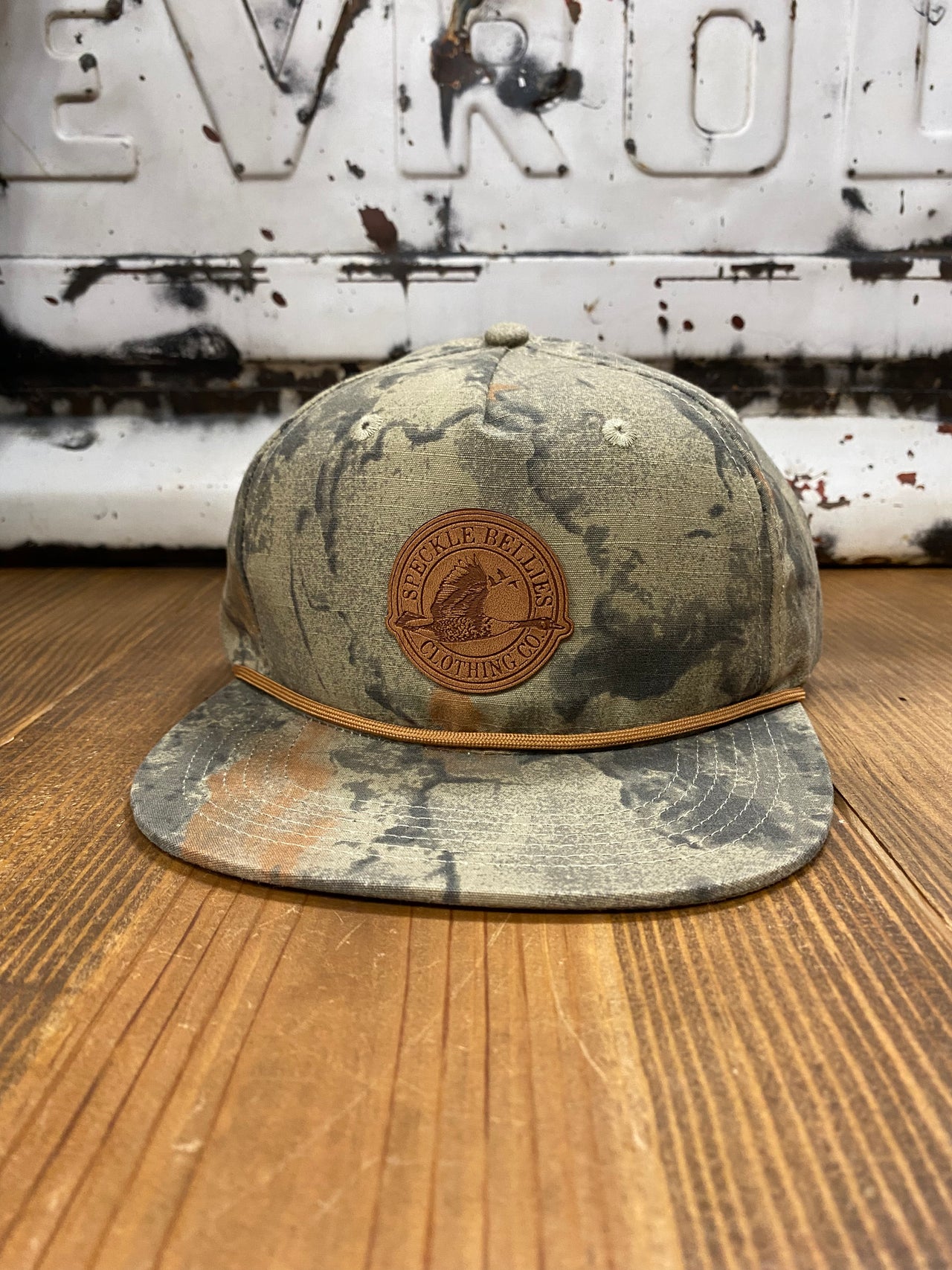 "Speckle Bellies Circle Logo Leather Patch Natural Camo Rope Cap - High-quality mesh and 100% polyester construction for comfort and durability. Features a distinctive leather patch with a circle logo, complemented by a natural camo pattern. Mid crown design, adjustable fit, and rugged rope detailing for an adventurous, outdoor-inspired look."