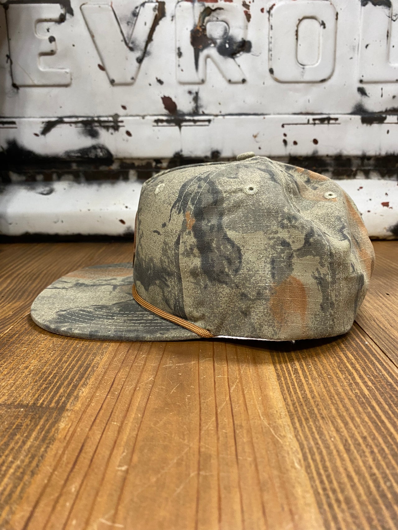 "Speckle Bellies Circle Logo Leather Patch Natural Camo Rope Cap - High-quality mesh and 100% polyester construction for comfort and durability. Features a distinctive leather patch with a circle logo, complemented by a natural camo pattern. Mid crown design, adjustable fit, and rugged rope detailing for an adventurous, outdoor-inspired look."